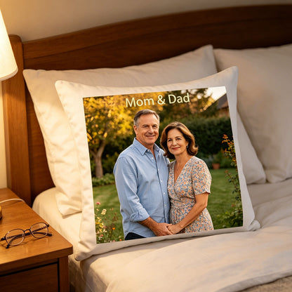 Custom Mom & Dad keepsake pillow resting on a sunlit bedroom bed, next to a nightstand with reading glasses. A tender, comforting scene showing the pillow as a sweet reminder of parents in a daughter’s personal space.